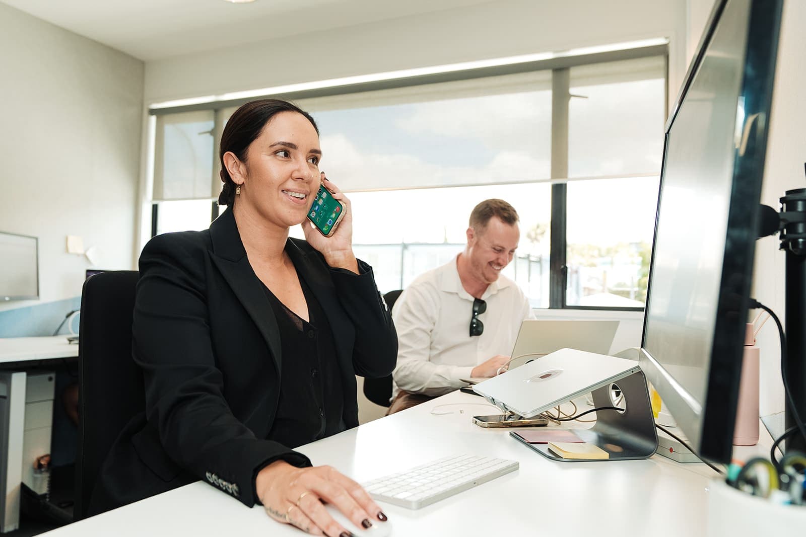 a professional lady in office space setting sitting on the desk, talking on her phone and looking at the screen