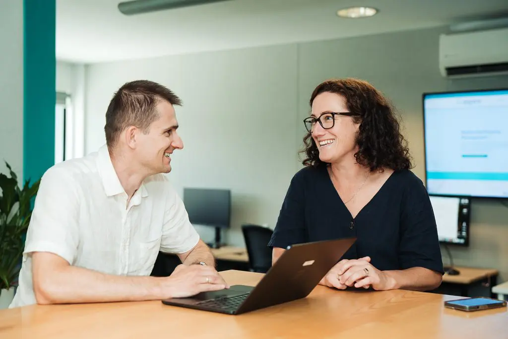 Two professionals in an office setting, one using a laptop while the other looks on and smiles, representing a collaborative property management team.